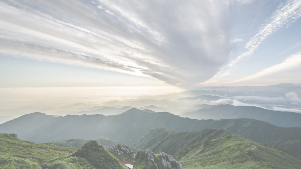 A panoramic view of misty mountains under a soft, cloudy sky during sunrise.